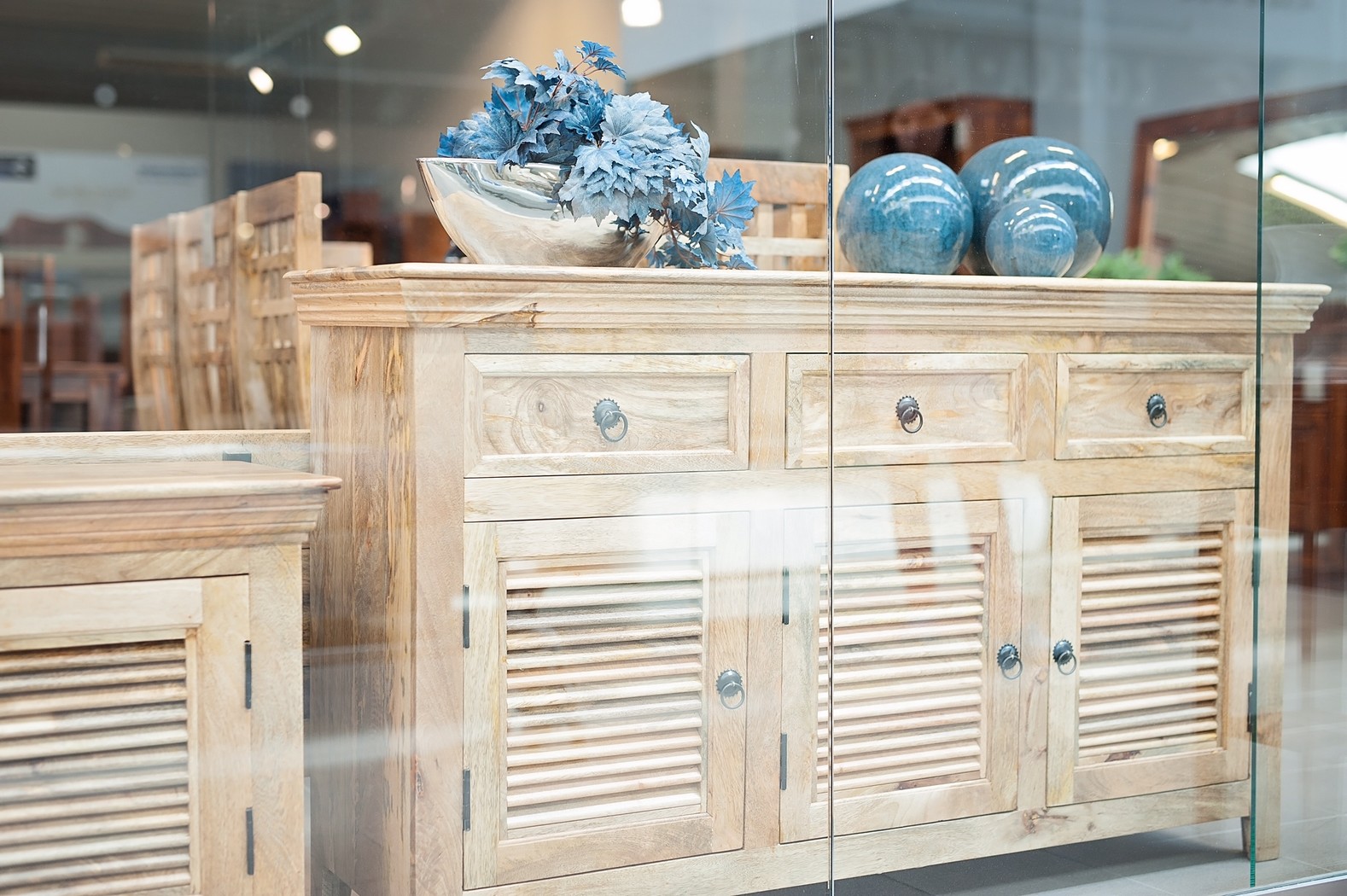 Jasny drewniany kredens z szufladami i żaluzjowymi drzwiczkami, udekorowany srebrną misą z niebieskimi kwiatami i trzema ceramicznymi kulami w odcieniach błękitu, widziany przez szybę wystawową.
