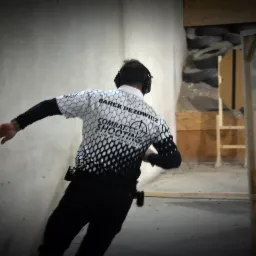 Mężczyzna w koszulce z napisem 'Combat Shooting' biegnie w pomieszczeniu strzeleckim z przeszkodami, widoczne ochraniacze na uszy i kabura z bronią przy pasie.