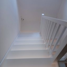 Pracownia Drzewna Sylwia Andrzejczyk - Minimalistyczne, białe schody z drewnianą balustradą, oświetlone od dołu, widziane z góry. Ujęcie podkreśla czystość formy i nowoczesny design.