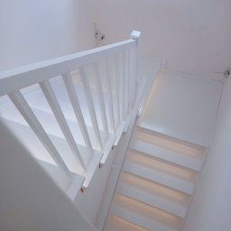Pracownia Drzewna Sylwia Andrzejczyk - Białe schody z drewnianą balustradą, widok z góry, oświetlone stopnie, minimalistyczny design, kabel wystający ze ściany, czyste, białe wnętrze.
