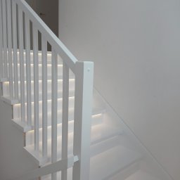Pracownia Drzewna Sylwia Andrzejczyk - Minimalistyczne, białe schody z podświetleniem LED, widok z dołu. Nowoczesny design, proste linie, jasne wnętrze. Neutralne tło, brak dekoracji.