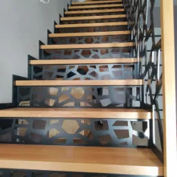Nowoczesne schody z drewnianymi stopniami i metalową balustradą o geometrycznym wzorze, widok z dołu.
