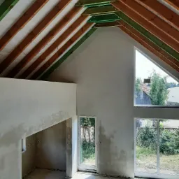 Surowe wnętrze domu w trakcie prac wykończeniowych: widoczne drewniane elementy konstrukcji dachu pomalowane na zielono i brązowo, ściany pokryte świeżym, szarym tynkiem, okna o nietypowym...