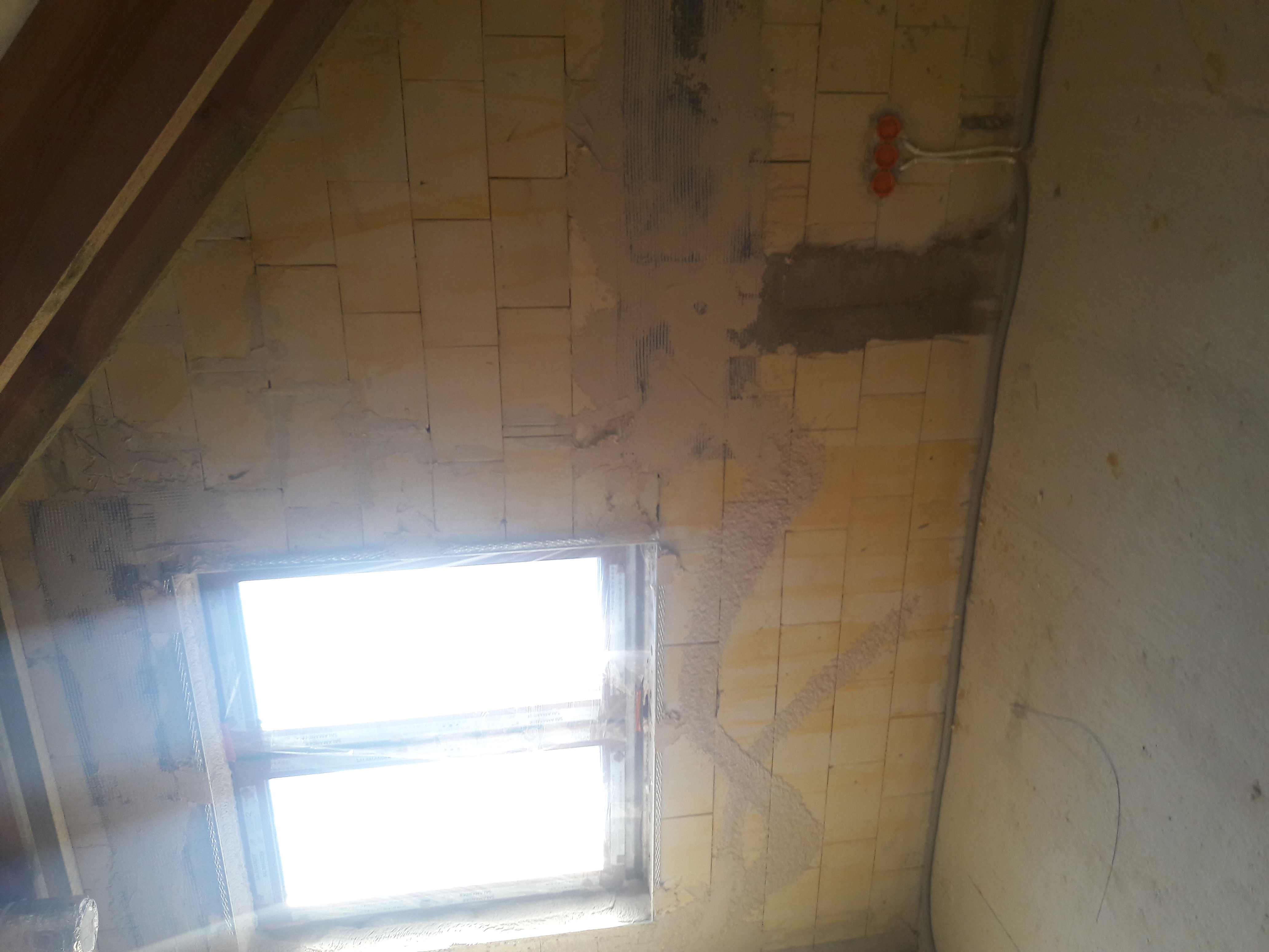 Ściana poddasza z oknem, częściowo pokryta siatką i zaprawą, przygotowana do nałożenia tynku maszynowego. Widoczne kable elektryczne w peszlach.