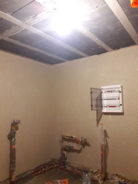 Świeżo położone, surowe ściany w Wołowie po aplikacji, widoczne wystające rury instalacji wodnej i otwarta skrzynka elektryczna, sufit z betonowych płyt.