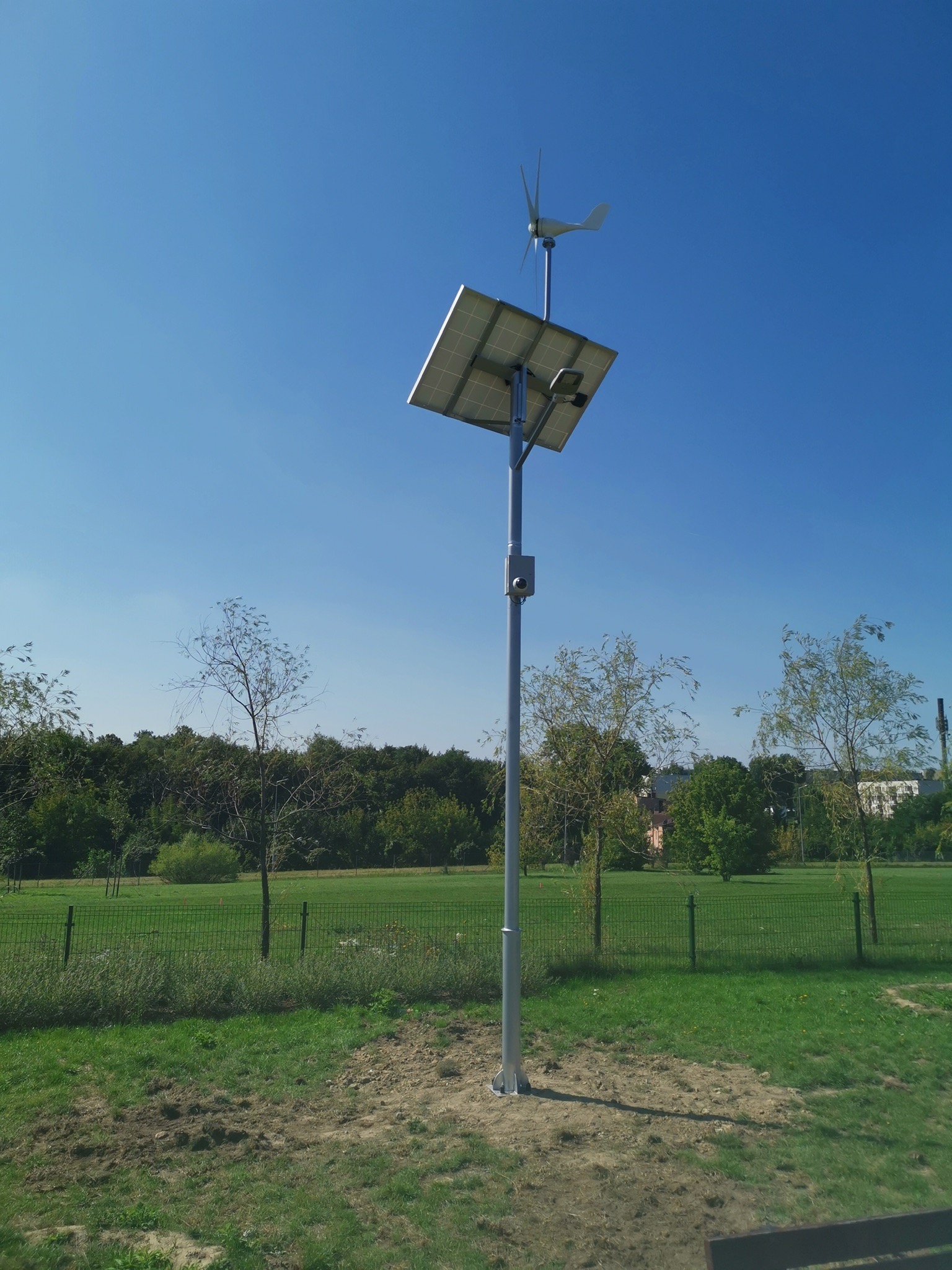 Słup oświetleniowy z panelem solarnym i turbiną wiatrową w parku, z widoczną kamerą monitoringu na słupie, na tle błękitnego nieba i zieleni drzew.