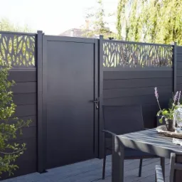 Nowoczesna, antracytowa furtka z ażurowym panelem górnym, wkomponowana w ogrodzenie z desek kompozytowych, widoczna z perspektywy tarasu z meblami ogrodowymi.