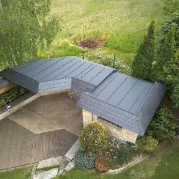 Widok z góry na budynek z dachem pokrytym szarą dachówką, przylegający do drewnianego tarasu i otoczony zielenią, z widoczną konstrukcją zadaszenia nad tarasem.