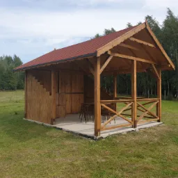 Drewniana konstrukcja z czerwonym dachem, częściowo zabudowana, z drewnianymi krzesłami i stołem w środku, na betonowej wylewce, otoczona trawnikiem i drzewami.