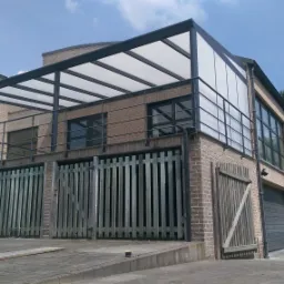 Dwukondygnacyjny budynek z cegły z metalową pergolą z białym zadaszeniem nad tarasem, drewniane ogrodzenie z pionowych desek, brama garażowa i samochód dostawczy w tle.