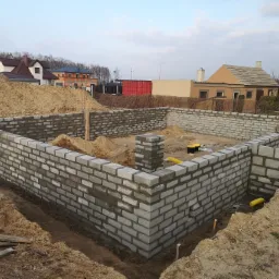 Wznoszenie fundamentów z bloczków betonowych; widoczne ułożone ściany fundamentowe z pustaków, zasypane ziemią z zewnątrz, z żółtymi rurami wewnątrz oraz stertą piasku w środku.