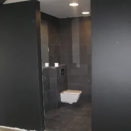 Nowoczesna łazienka z szarymi płytkami i podwieszaną toaletą, widok zza czarnej ściany działowej, dwie rolki papieru toaletowego na półce.