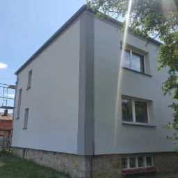 Dwukondygnacyjny dom z jasnoszarą elewacją i kamienną podmurówką, widoczny z boku, z rusztowaniem w tle i drzewem po prawej stronie kadru, w słoneczny dzień.