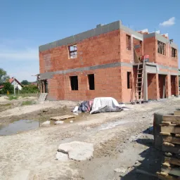 Dwukondygnacyjny budynek w trakcie wznoszenia z czerwonej cegły, widoczne niezamurowane otwory okienne, drewniana drabina oparta o ścianę, teren budowy z błotem i kałużami.