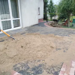 W trakcie budowy tarasu: sterta piasku na częściowo ułożonej kostce brukowej z czerwonym obramowaniem, szpadel wbity w piasek, dom z dużymi oknami w tle.