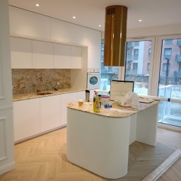 Daniel Szymańczak - Nowoczesna kuchnia w trakcie wykańczania, z białymi szafkami, blatem z wzorem kamienia, złotą armaturą i okrągłym wyspowym blatem, widoczne materiały budowlane i środki czystości na blacie, podłoga...