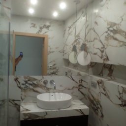 Daniel Szymańczak - Nowoczesna łazienka z marmurowymi płytkami na ścianach i blacie, okrągła biała umywalka nablatowa, wiszące lampy z okrągłymi kloszami i widoczne odbicie osoby robiącej zdjęcie w lustrze.