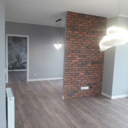 Daniel Szymańczak - Salon po remoncie z drewnianą podłogą, szarą ścianą, ścianką działową z cegły klinkierowej i widokiem na pokój z tapetą w motywy roślinne.