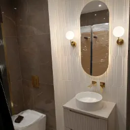 Nowoczesna łazienka z marmurową ścianą, białą umywalką nablatową z szafką, podświetlanym owalnym lustrem i złotą armaturą. Toaleta w folii ochronnej z portfelem na wierzchu.