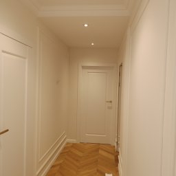 Daniel Szymańczak - Korytarz w jasnych barwach po remoncie, z parkietem ułożonym we wzór jodełki, ozdobnymi listwami ściennymi i trzema punktowymi lampami w suficie.
