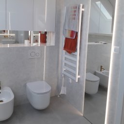 Daniel Szymańczak - Nowoczesna łazienka w odcieniach szarości z białymi sanitariatami, podświetlanym lustrem i grzejnikiem drabinkowym z ręcznikami w kolorach ziemi.