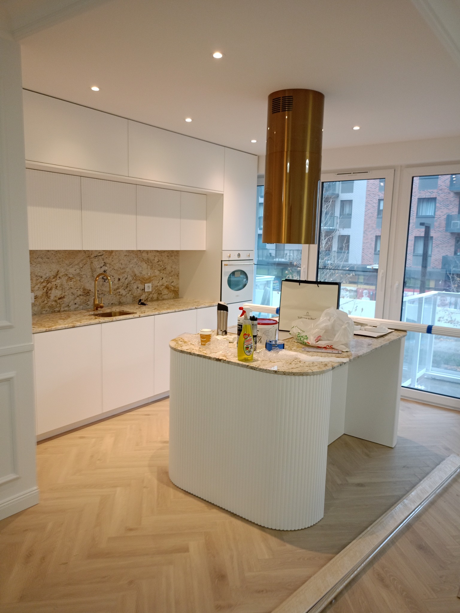 Nowoczesna kuchnia w trakcie wykańczania, z białymi szafkami, blatem z wzorem kamienia, złotą armaturą i okrągłym wyspowym blatem, widoczne materiały budowlane i środki czystości na blacie, podłoga...