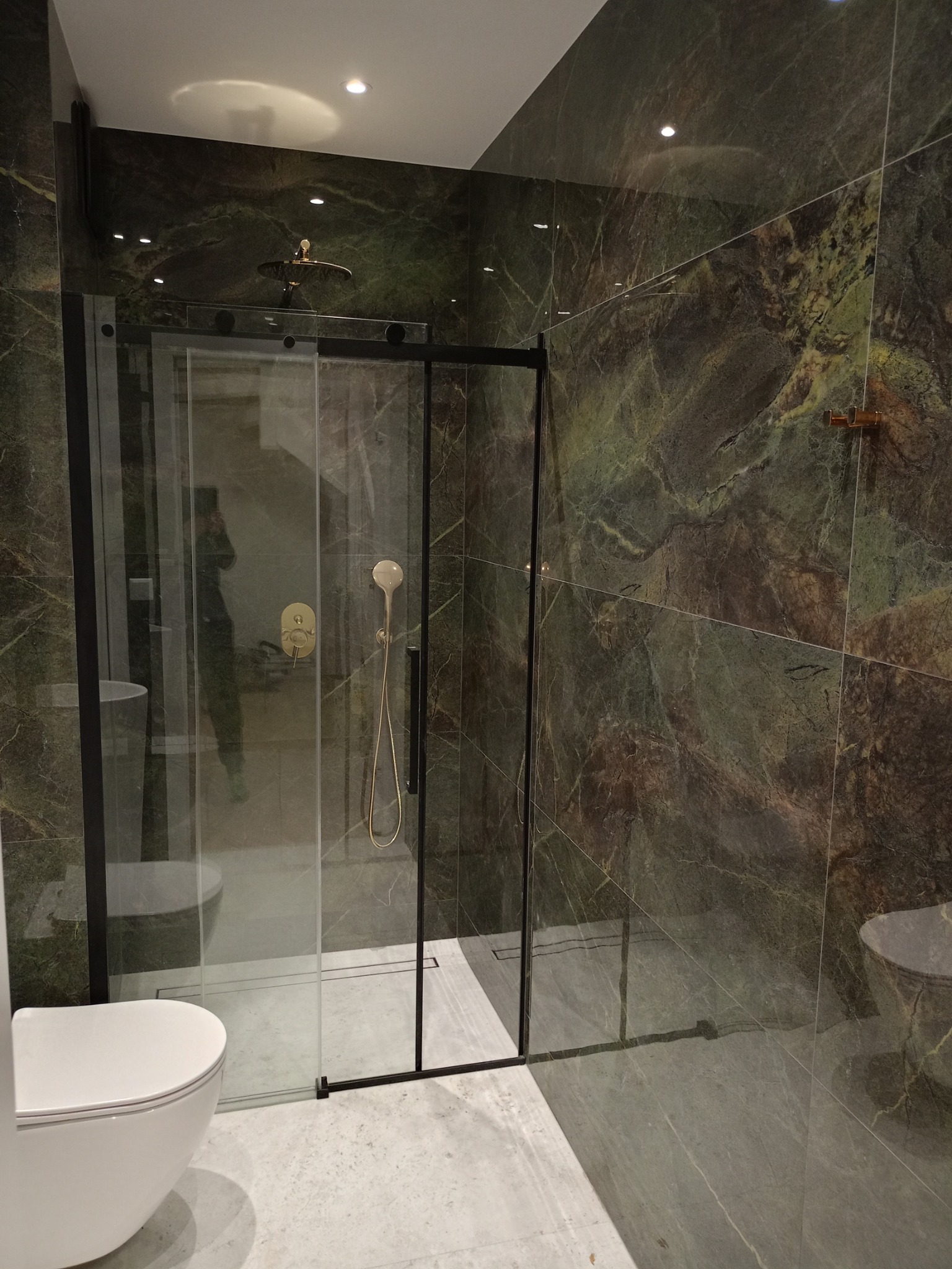 Nowoczesna łazienka z kabiną prysznicową typu walk-in z czarnymi ramami, armaturą w kolorze szczotkowanego złota i płytkami imitującymi zielony marmur na ścianach i podłodze, widok na toaletę.