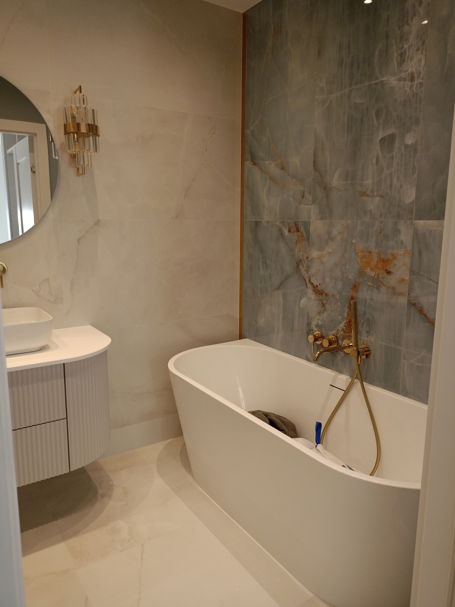 Nowoczesna łazienka z wolnostojącą wanną, marmurowymi płytkami w odcieniach szarości i złota armaturą. Okrągłe lustro nad umywalką i kryształowy kinkiet dodają elegancji.