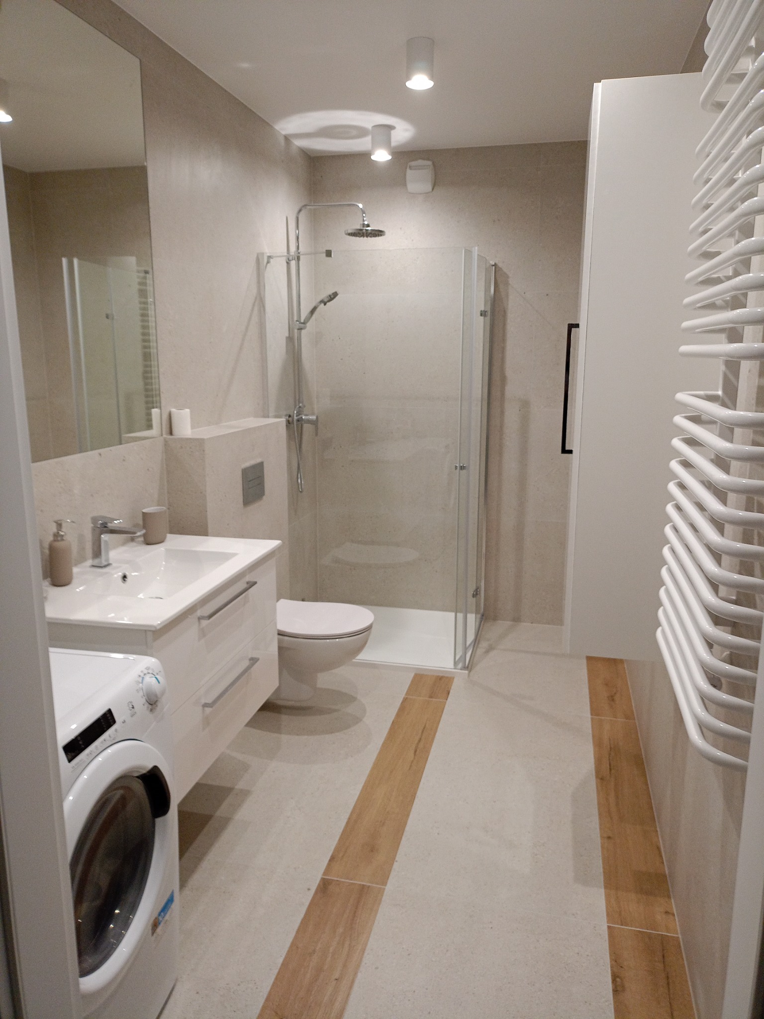 Jasna łazienka z kabiną prysznicową, umywalką z szafką, pralką i grzejnikiem drabinkowym; podłoga wyłożona płytkami w odcieniach beżu z drewnopodobnymi wstawkami.