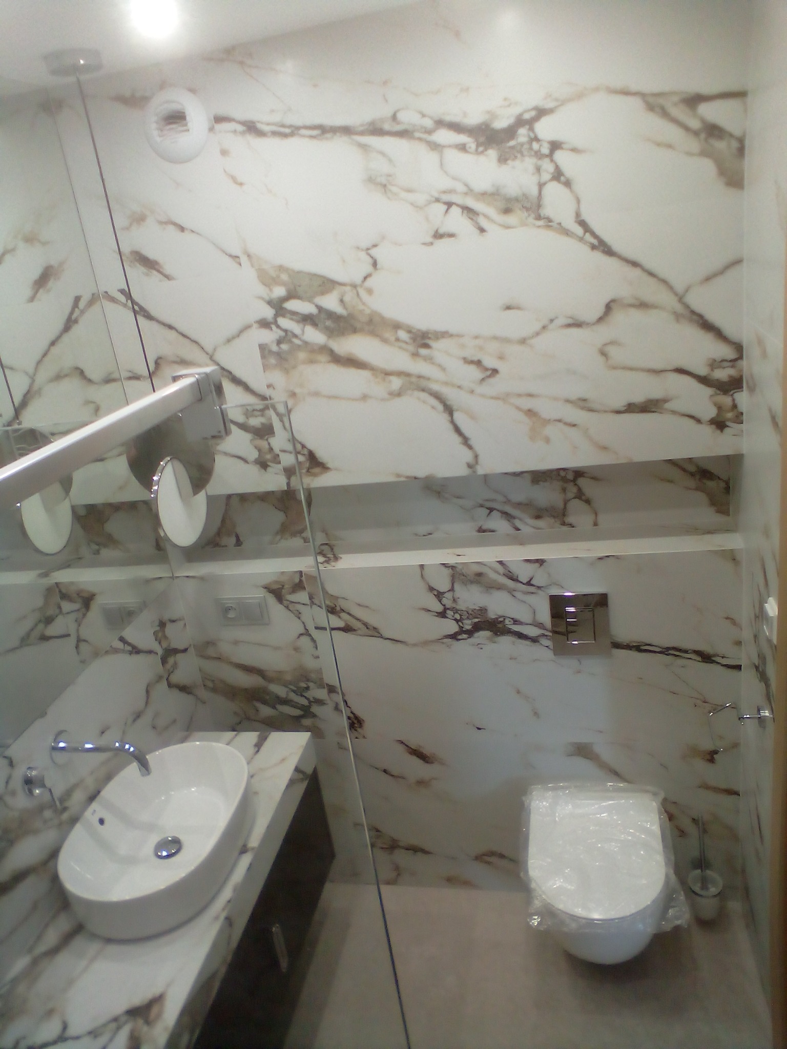 Wykończona łazienka z marmurowymi płytami na ścianach, umywalka nablatowa na blacie z tego samego marmuru, podwieszana toaleta z plastikową folią ochronną na desce, oświetlenie punktowe sufitowe.