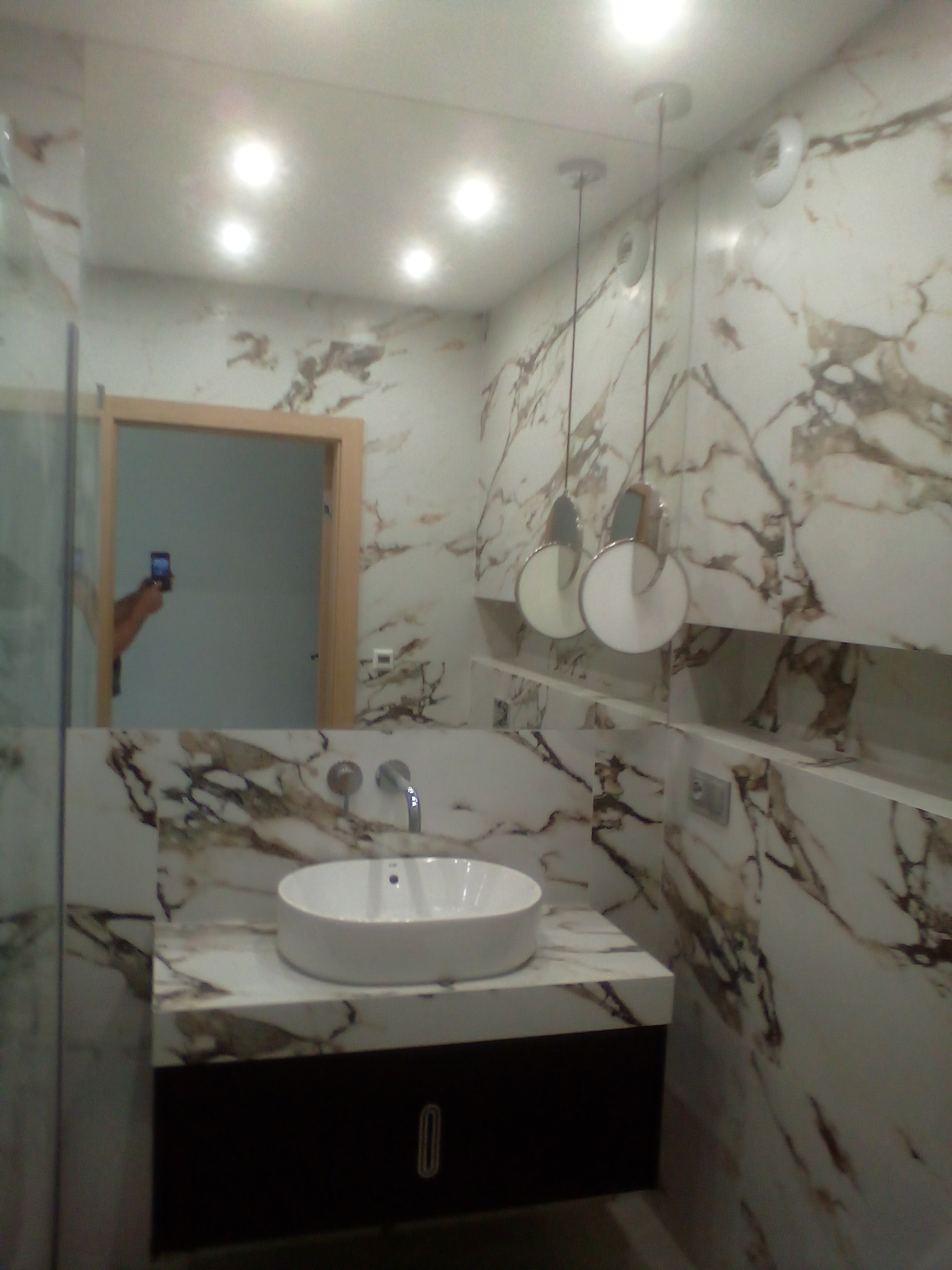 Nowoczesna łazienka z marmurowymi płytkami na ścianach i blacie, okrągła biała umywalka nablatowa, wiszące lampy z okrągłymi kloszami i widoczne odbicie osoby robiącej zdjęcie w lustrze.