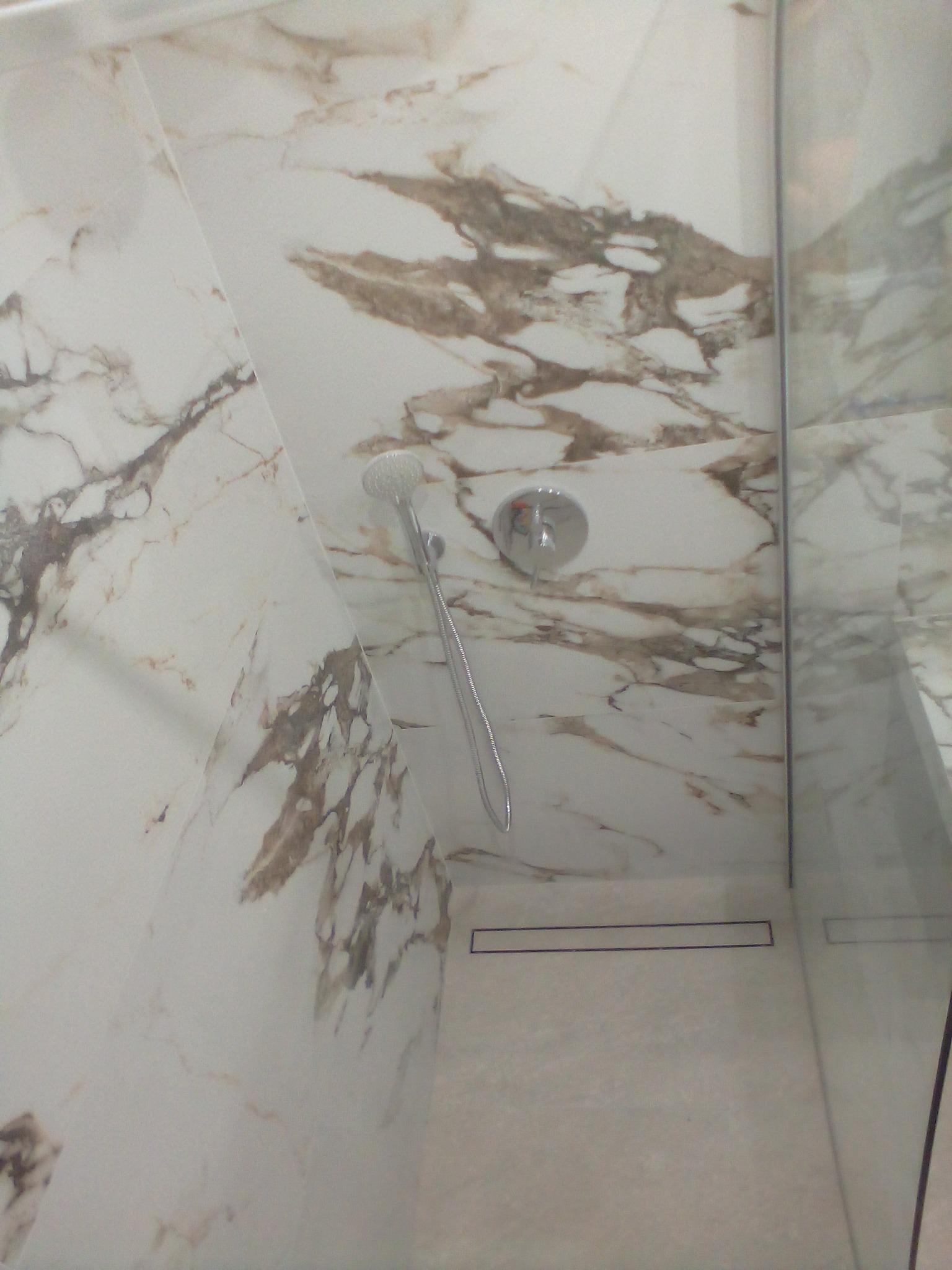 Kabina prysznicowa wykończona białymi płytami imitującymi marmur z brązowymi żyłami, widoczna deszczownica i odpływ liniowy