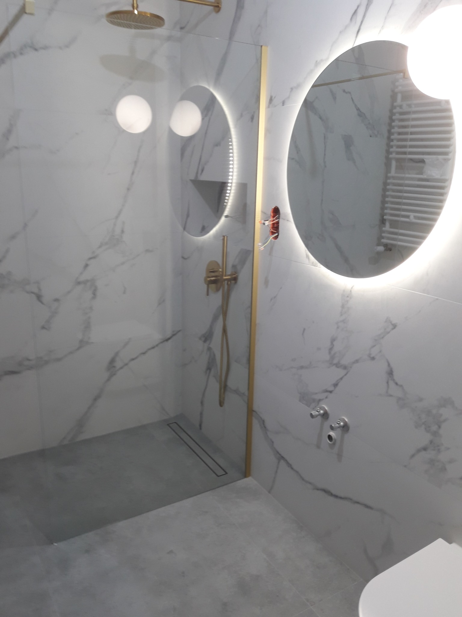 Nowoczesna łazienka z marmurowymi płytkami, złotą armaturą, oświetlonym okrągłym lustrem i szarą strefą prysznicową z odpływem liniowym.