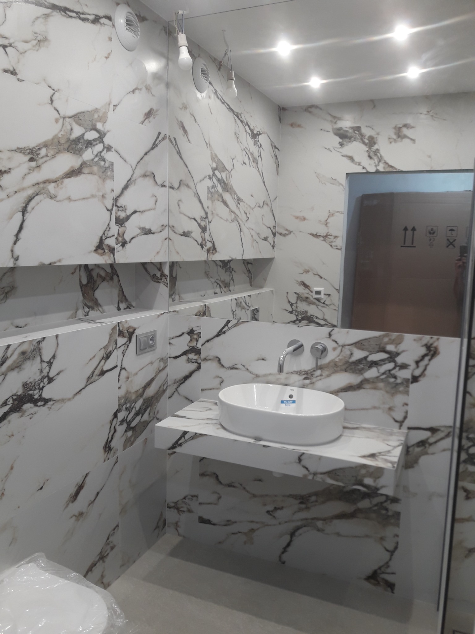 Nowoczesna łazienka z marmurowymi płytkami, okrągła umywalka nablatowa na blacie z tego samego materiału, bateria ścienna, widoczne elementy instalacji elektrycznej i wentylacji.