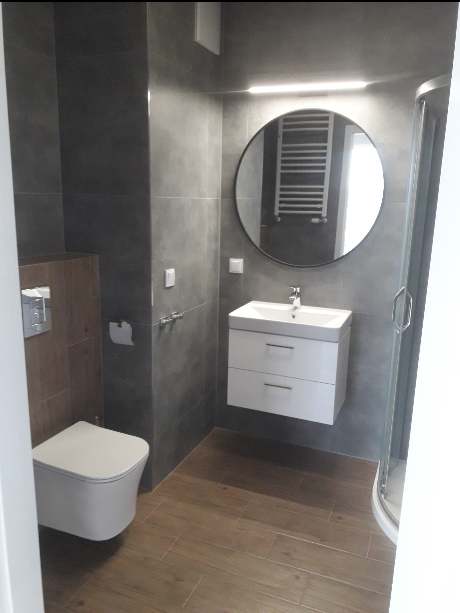 Nowoczesna łazienka z szarymi płytkami, drewnopodobną podłogą, okrągłym lustrem z podświetleniem LED i wiszącą szafką z umywalką.