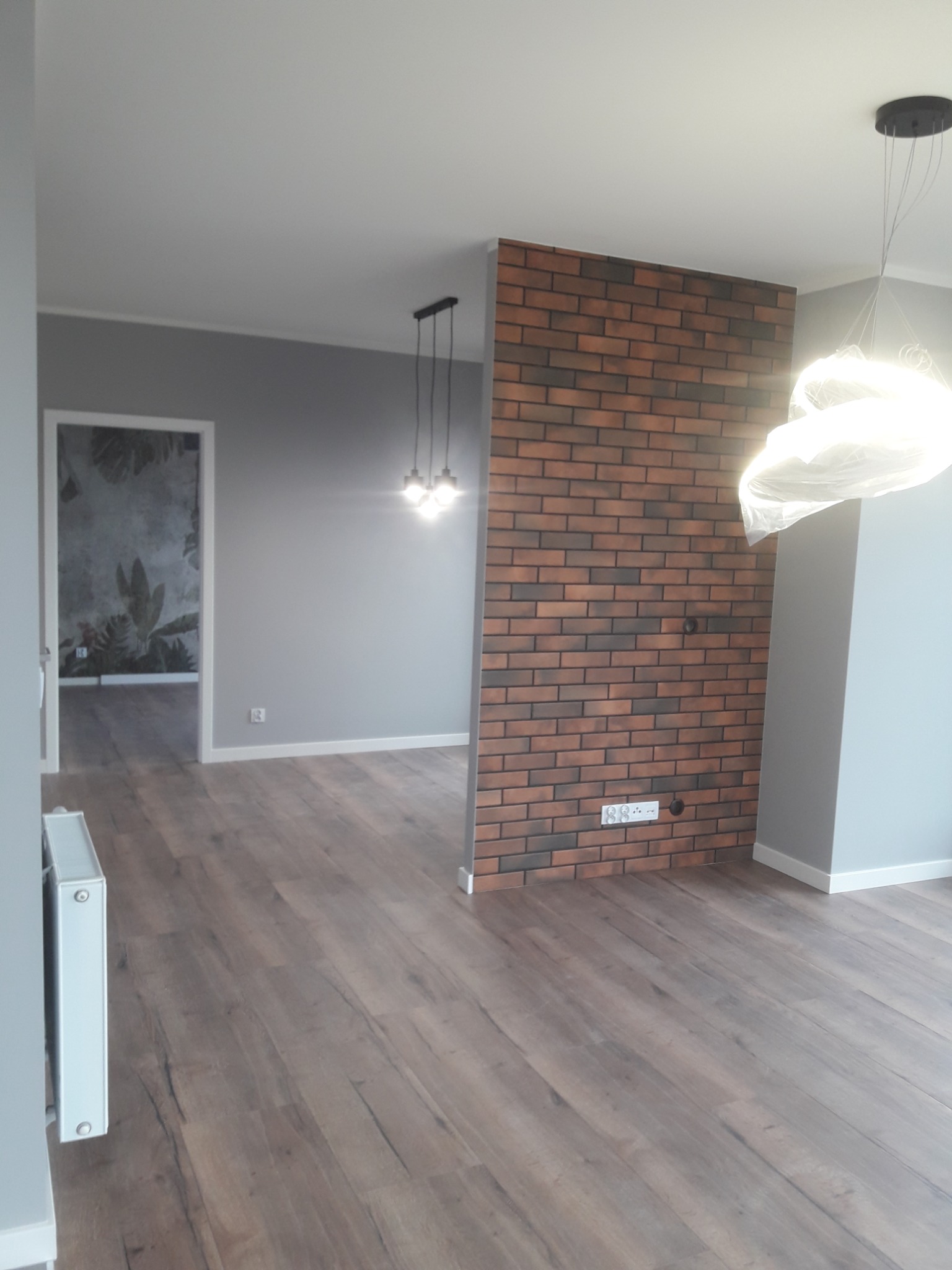 Salon po remoncie z drewnianą podłogą, szarą ścianą, ścianką działową z cegły klinkierowej i widokiem na pokój z tapetą w motywy roślinne.