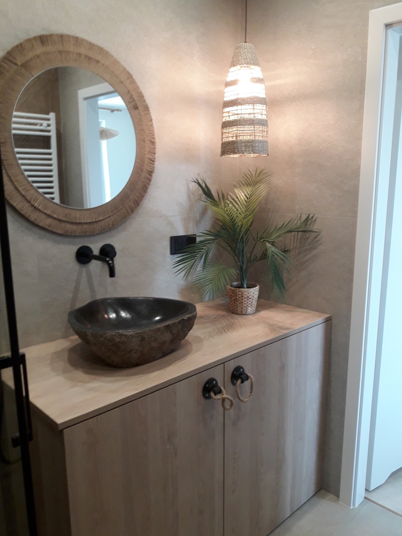 Nowoczesna łazienka z okrągłym lustrem w ramie z naturalnego sznurka, kamienną umywalką nablatową, szafką z drewnopodobnymi frontami i plecionymi uchwytami sznurkowymi oraz dekoracyjną lampą...