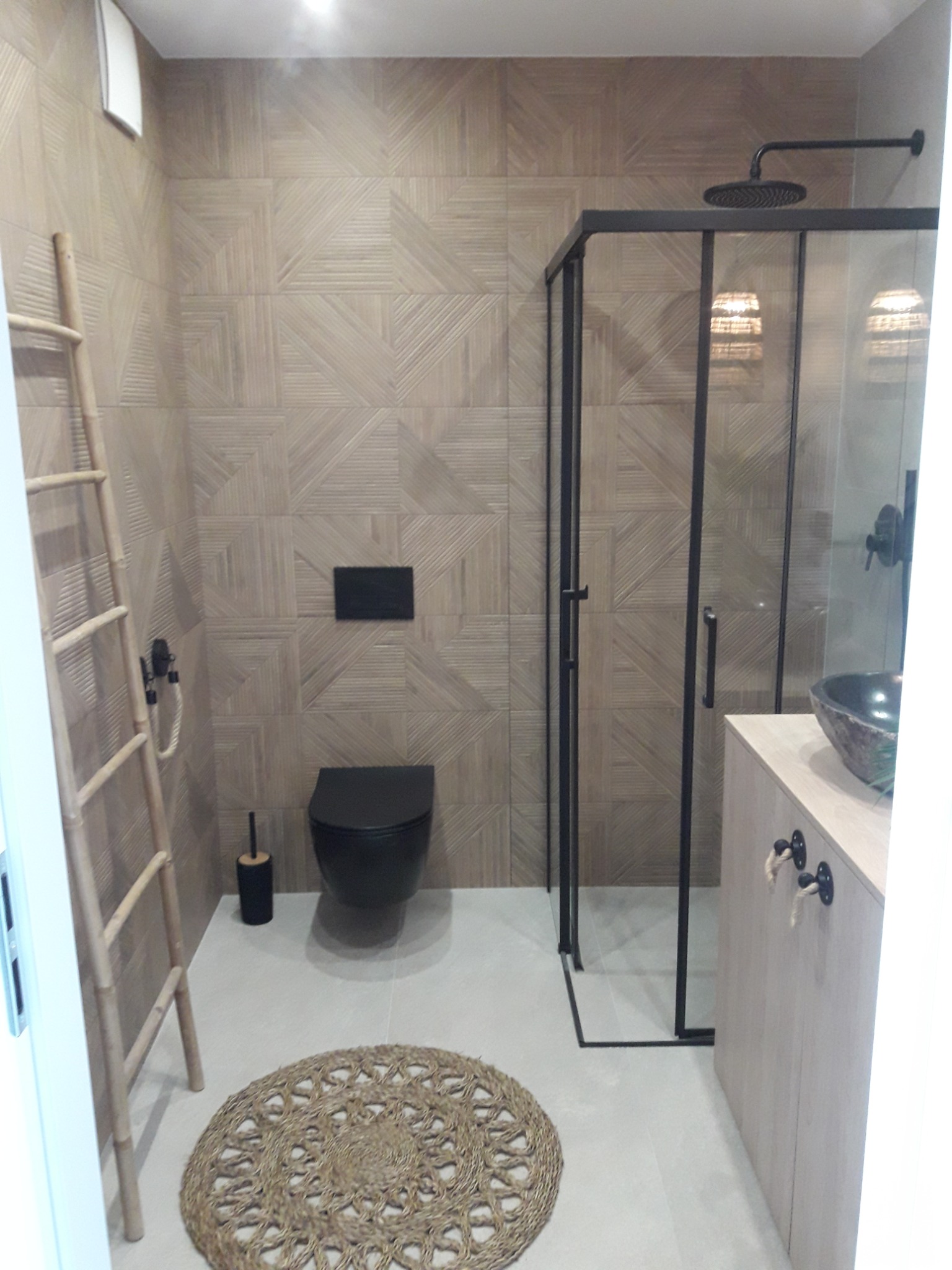 Nowoczesna łazienka z geometrycznymi płytkami w odcieniach beżu, czarną armaturą, kabiną prysznicową typu walk-in z czarnymi profilami, umywalką nablatową z kamienia i drabinką dekoracyjną.