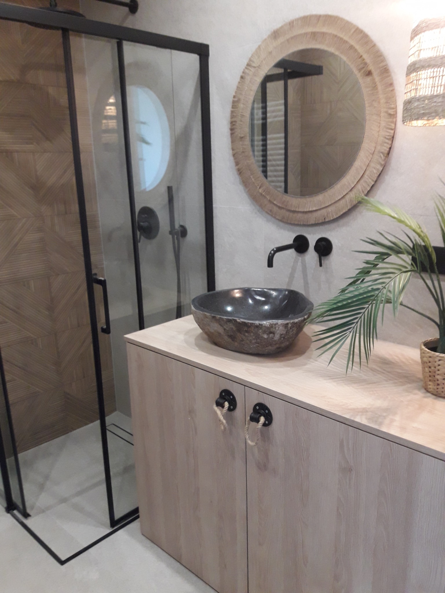 Nowoczesna łazienka z kabiną prysznicową z czarną ramą, okrągłym lustrem w naturalnej ramie, kamienną umywalką nablatową i szafką z jasnego drewna z nietypowymi uchwytami z liny.
