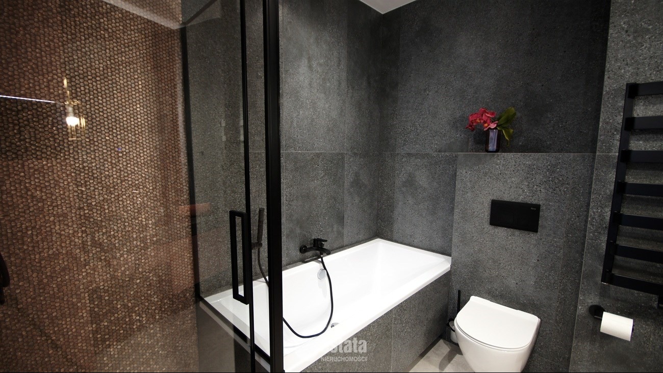 Nowoczesna łazienka z kabiną prysznicową, mozaiką w odcieniach brązu, szarą ścianą z kwiatami w wazonie, białą wanną i toaletą oraz czarnymi elementami armatury i grzejnika.