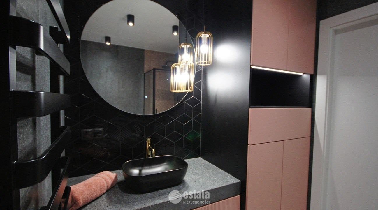 Nowoczesna łazienka z czarną umywalką nablatową, okrągłym lustrem z podświetleniem, złotymi lampami wiszącymi i szafkami w odcieniach różu i czerni, czarne płytki z geometrycznym wzorem na ścianie.