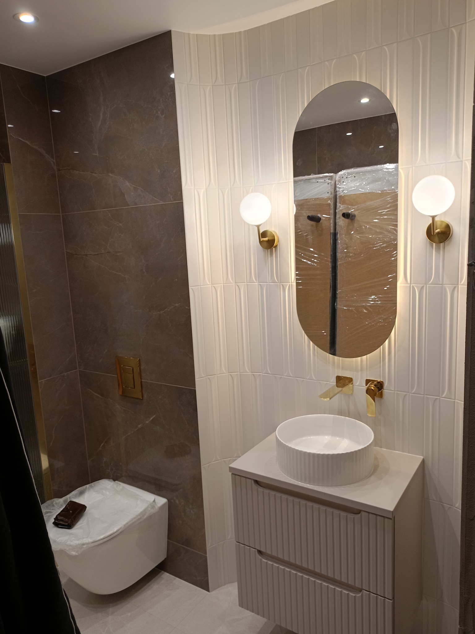 Nowoczesna łazienka z marmurową ścianą, białą umywalką nablatową z szafką, podświetlanym owalnym lustrem i złotą armaturą. Toaleta w folii ochronnej z portfelem na wierzchu.