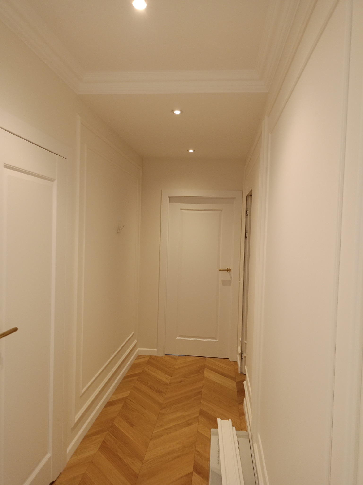 Korytarz w jasnych barwach po remoncie, z parkietem ułożonym we wzór jodełki, ozdobnymi listwami ściennymi i trzema punktowymi lampami w suficie.