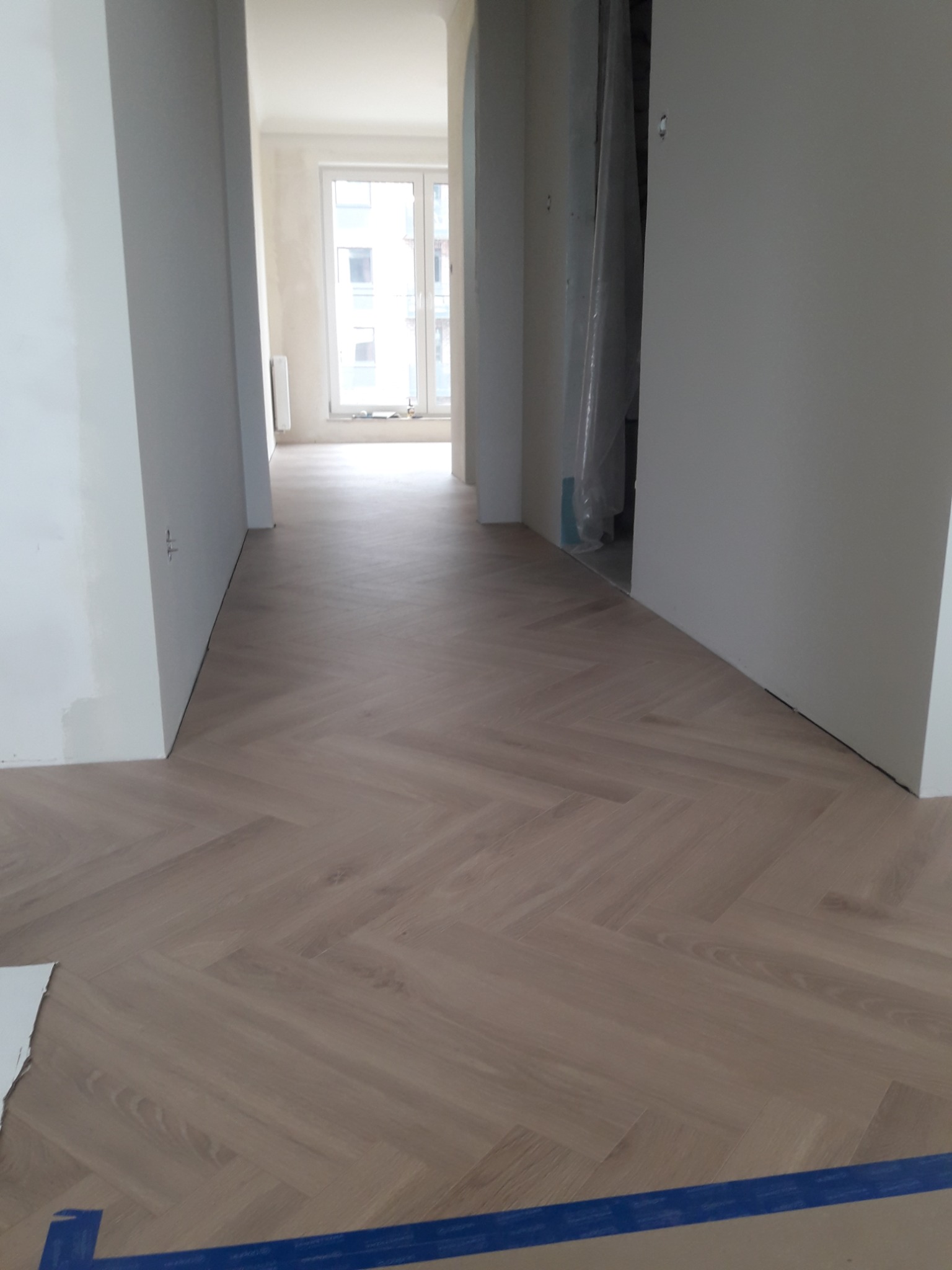 Nowo ułożona, jasna podłoga z paneli w jodełkę w korytarzu mieszkania w trakcie wykańczania, zabezpieczona niebieską taśmą malarską przy krawędziach. Widoczne surowe ściany i framugi drzwi.