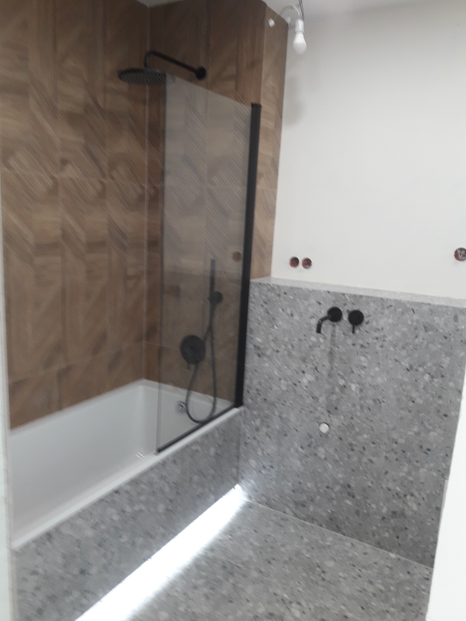 Nowoczesna łazienka z drewnopodobnymi płytkami ułożonymi w jodełkę, kabiną prysznicową z czarną ramą i deszczownicą, szarą posadzką i ścianą z terazzo oraz czarną armaturą. Widoczne niedokończone...