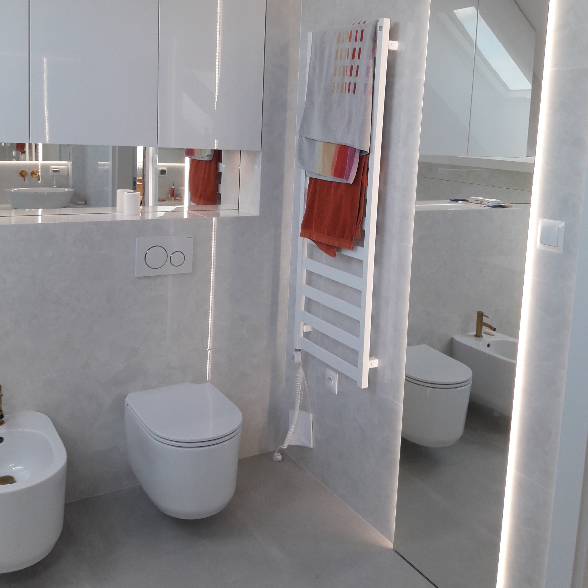 Nowoczesna łazienka w odcieniach szarości z białymi sanitariatami, podświetlanym lustrem i grzejnikiem drabinkowym z ręcznikami w kolorach ziemi.