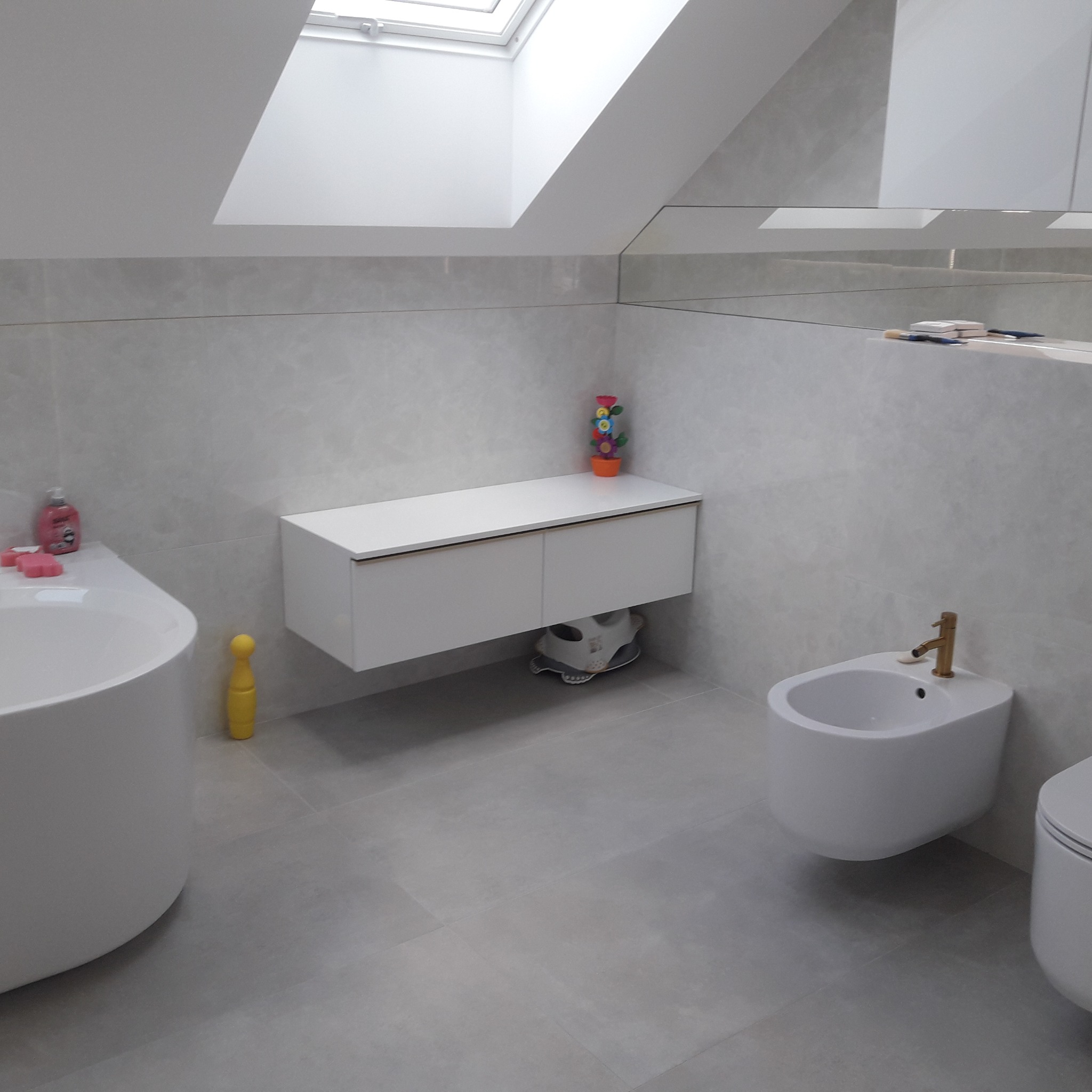 Nowoczesna łazienka na poddaszu z szarymi płytkami, owalną wanną, wiszącą szafką z umywalką, bidetem z złotą armaturą i oknem dachowym.