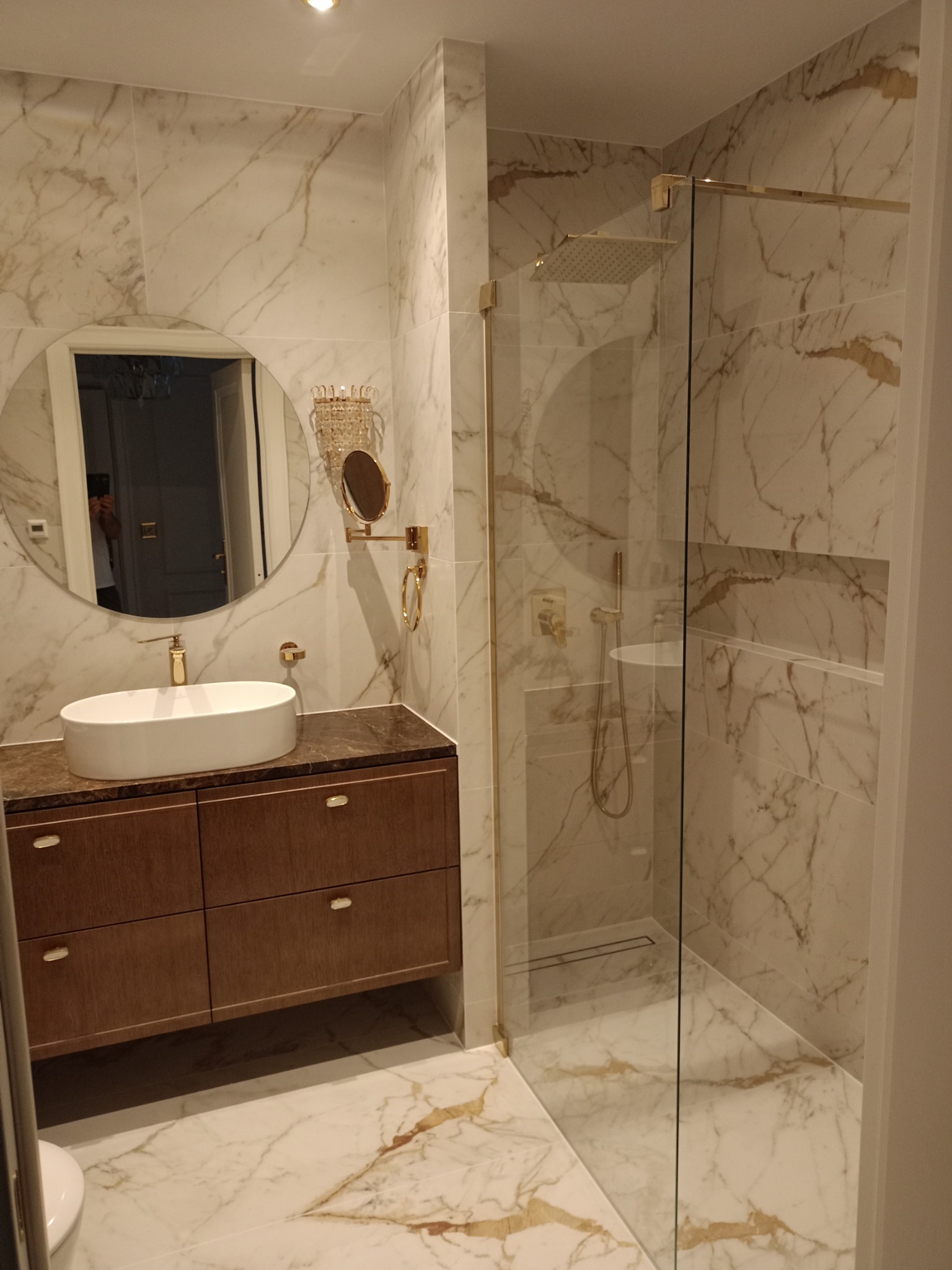 Elegancka łazienka z marmurowymi płytkami, kabiną prysznicową typu walk-in ze złotymi detalami, umywalką nablatową na szafce z drewnopodobnymi frontami i okrągłym lustrem z podświetleniem.