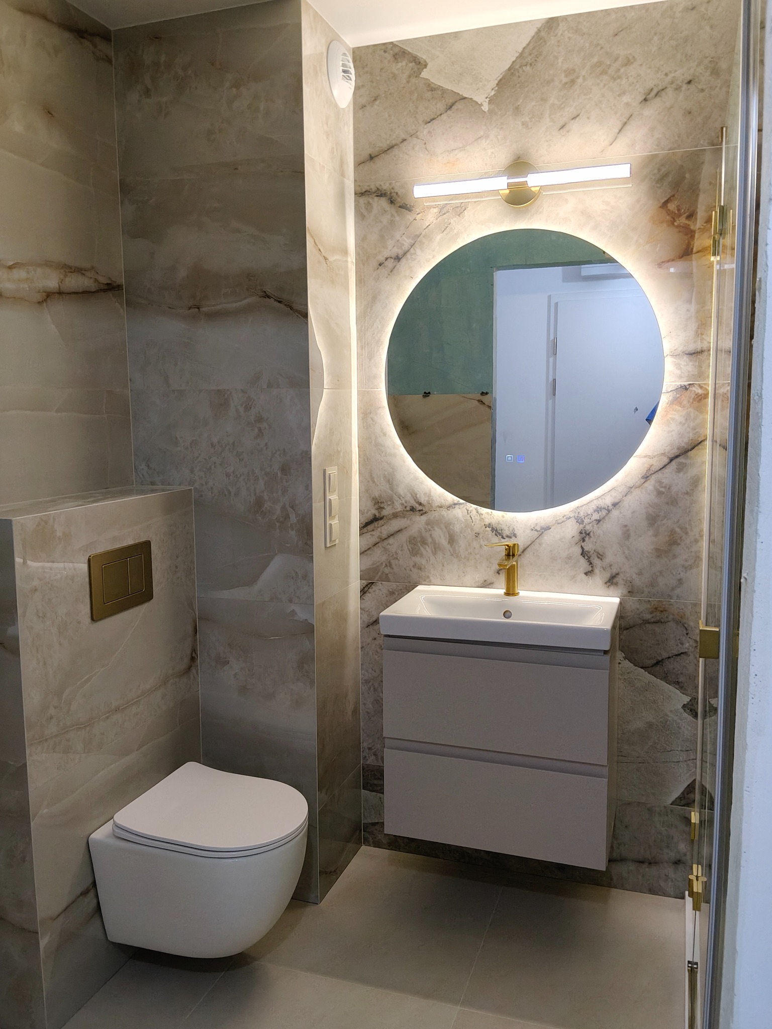 Elegancka łazienka z marmurowymi płytkami, podwieszaną toaletą i okrągłym lustrem z podświetleniem LED. Złote akcenty armatury i detali dodają luksusowego charakteru.