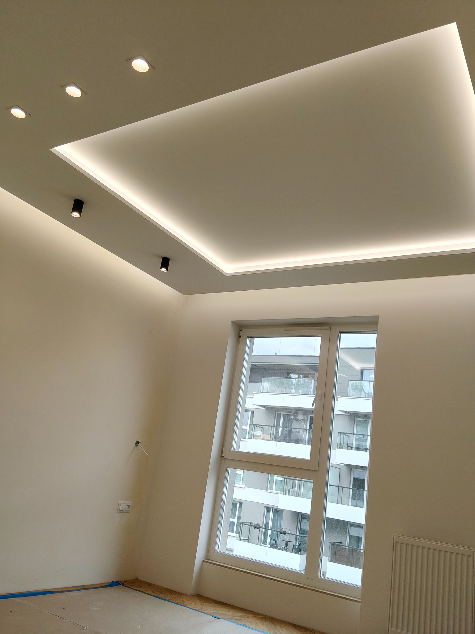 Nowoczesny sufit podwieszany z oświetleniem LED i punktowymi lampami w jasnym pokoju z oknem. Widok na budynek z balkonami.